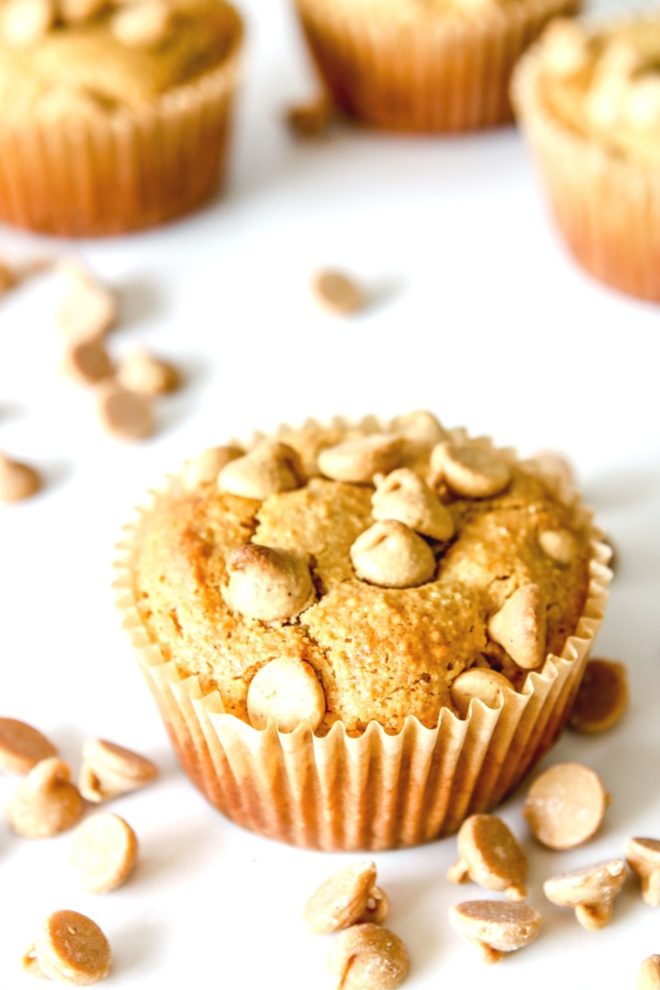 This is an overhead side angle view of a peanut butter muffin with peanut butter chips on top. The muffin sits on a white counter with more muffins blurred in the background. Peanut butter chips are scattered around the muffins.