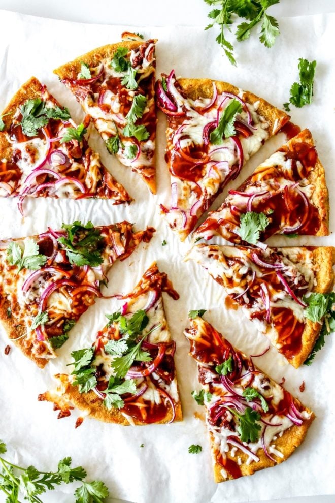 This is an overhead image of slices of pumpkin pizza topped with bbq sauce, cheese, red onion and cilantro. The pizza sits on a white pieces of parchment paper with cilantro surrounding it.