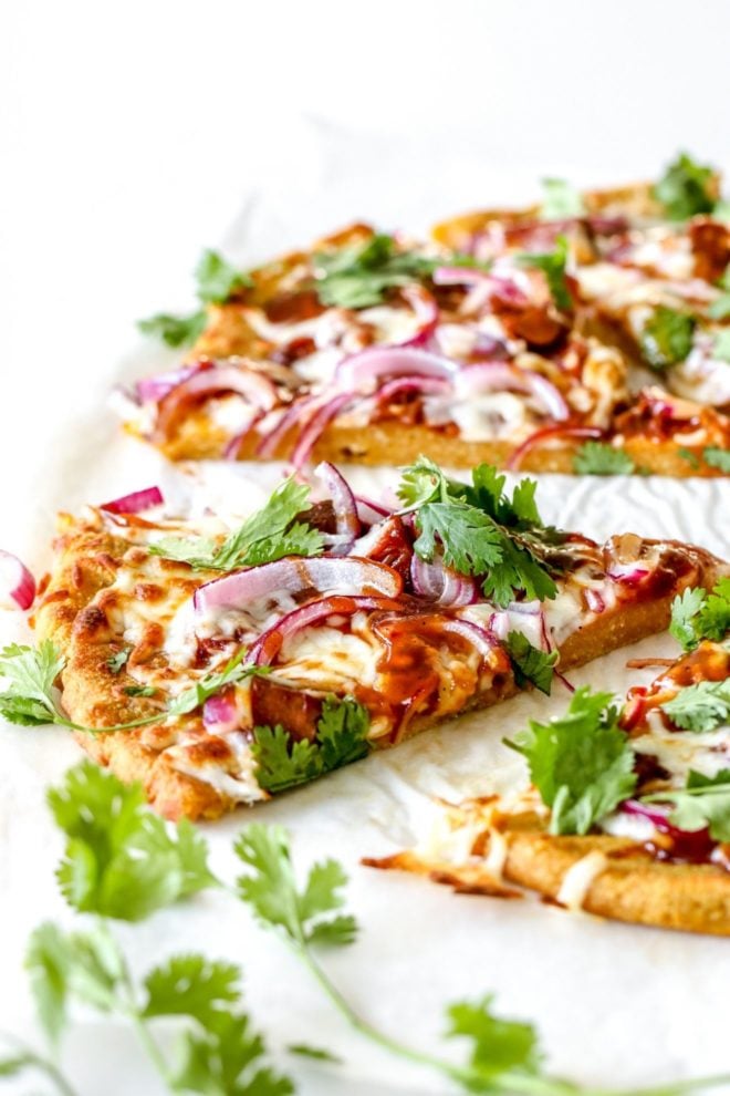 This is a side view of a slice of pumpkin pizza topped with bbq sauce, cheese, red onion, and cilantro. More pizza slices are blurred in the back. The slice sits on a white piece of parchment paper with cilantro leaves around it.