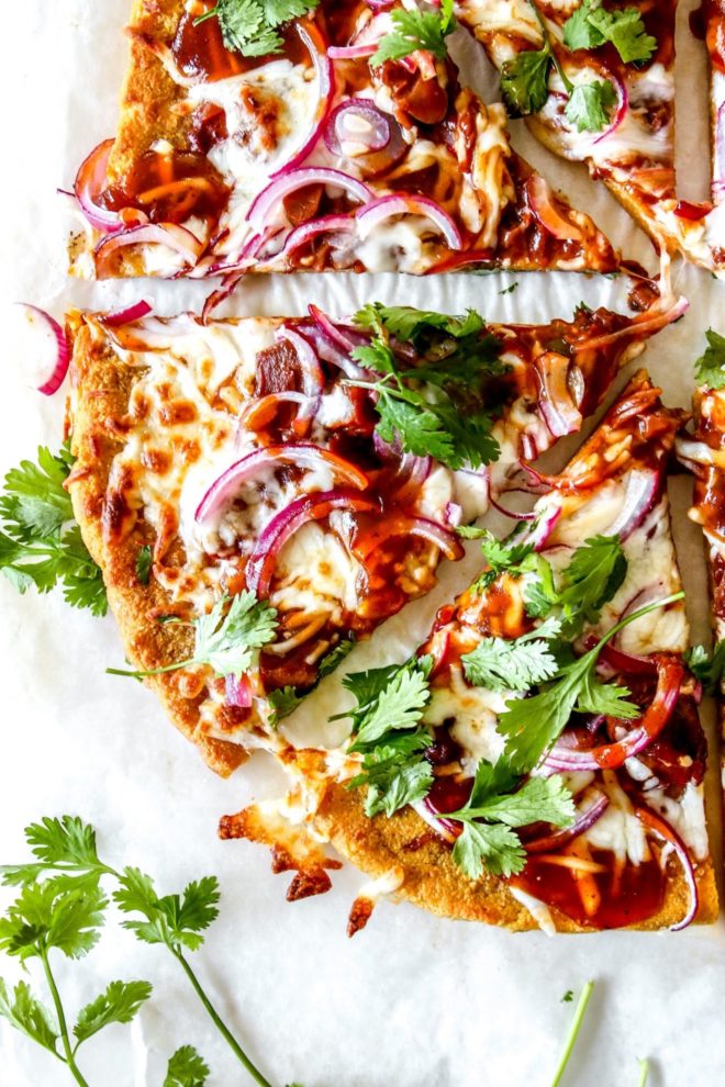 This is an overhead closeup of a slice of pumpkin pizza topped with red onions, cheese, bbq sauce and cilantro. The slices sit on a white piece of parchment paper.