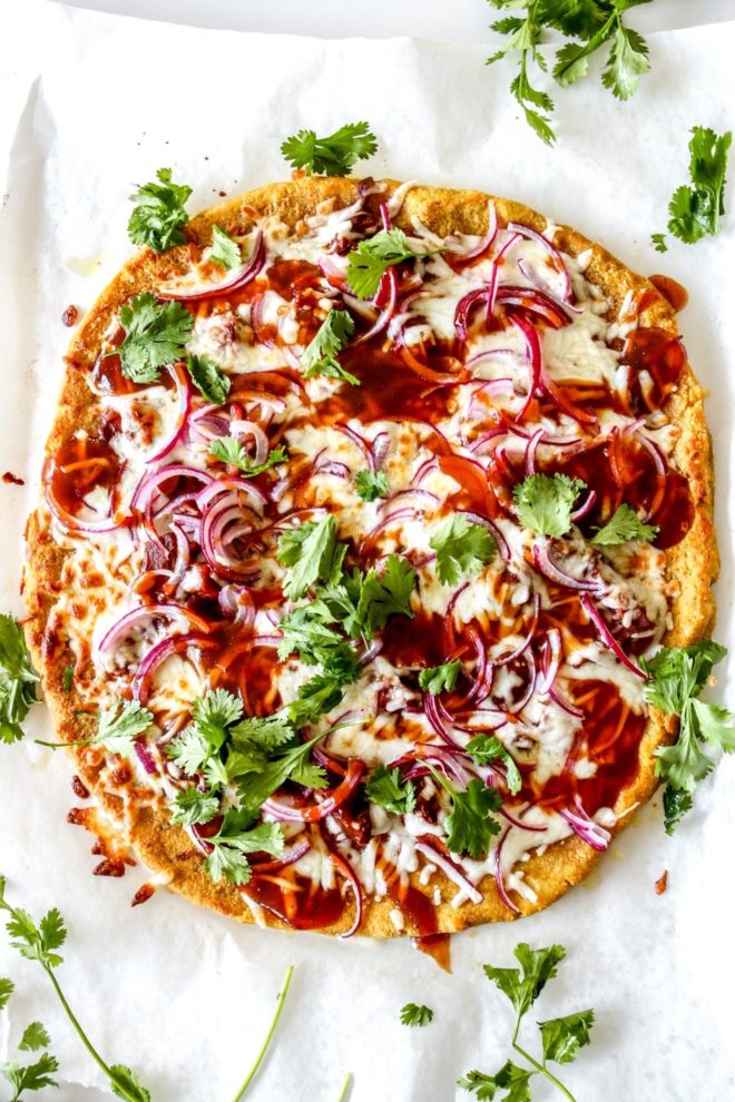 This is an overhead image of a pumpkin pizza topped with bbq sauce, melted cheese, red onion, and cilantro. The pizza sits on a white piece of parchment paper with more cilantro around the pizza.