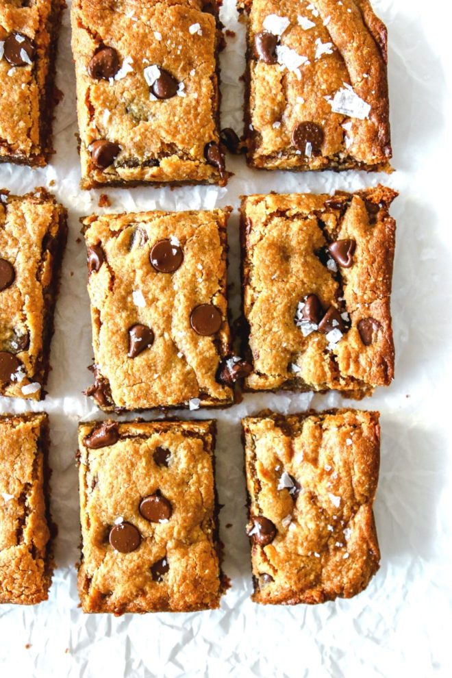 This is an overhead image of chocolate chip blondies sitting on a white piece of parchment paper. The blondies are sprinkled with flakey salt.