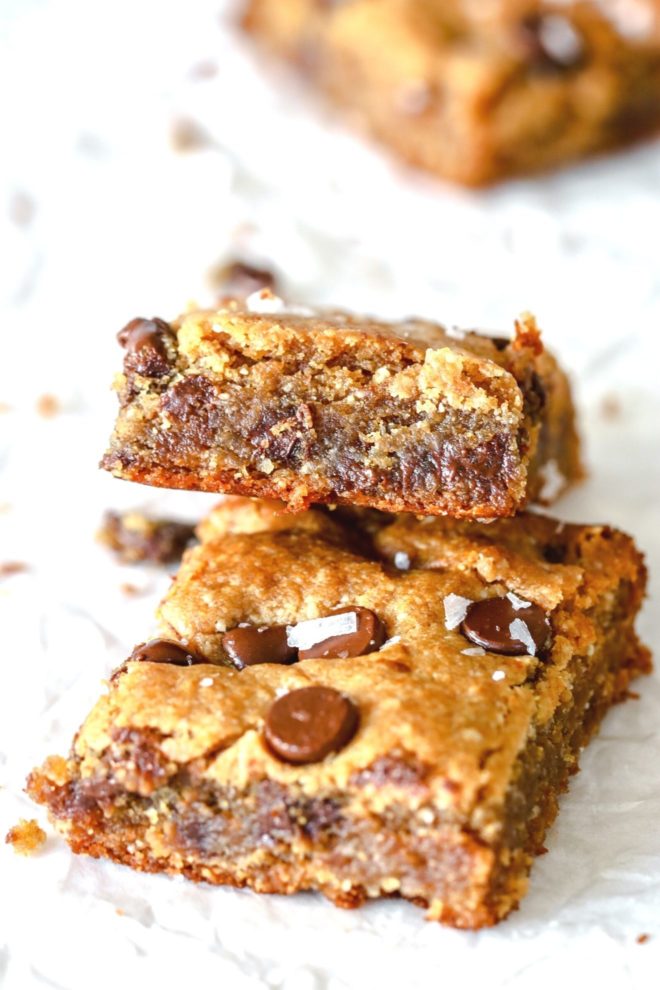 This is a side view of one chocolate chip blondie leaning against another. The blondies sit on a white surface with another blurred in the background.