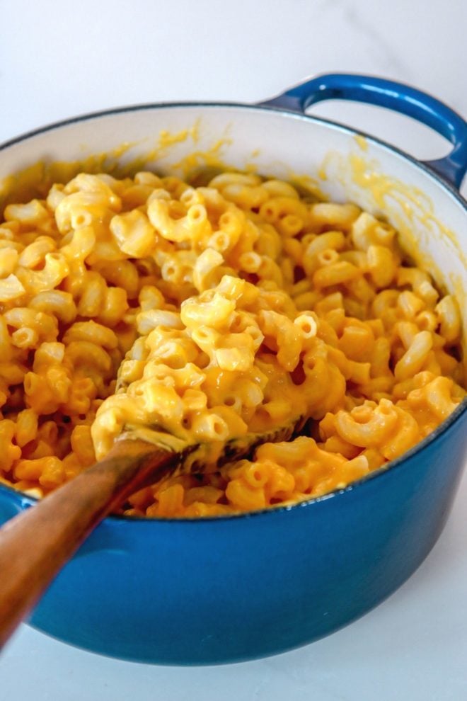 This is a side view of a blue pot filled with mac and cheese. A wooden spoon is scooping up some noodles and leaning against the side of the pot.