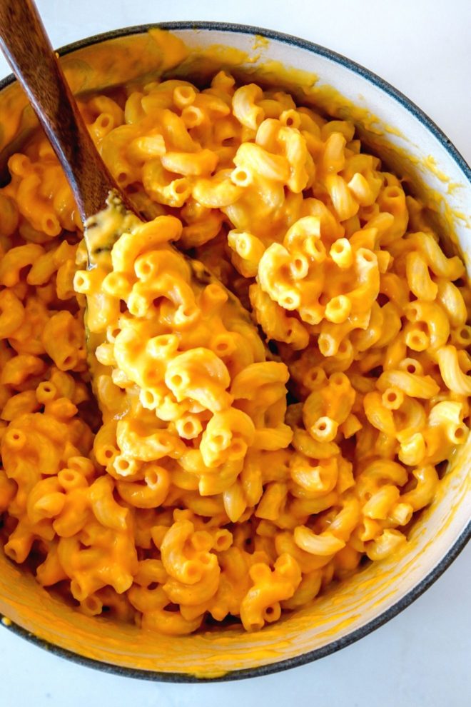 This is an overhead image of a pot filled with cheesy macaroni noodles. A wooden spoon is scooping up some noodles and leaning against the side of the pot.