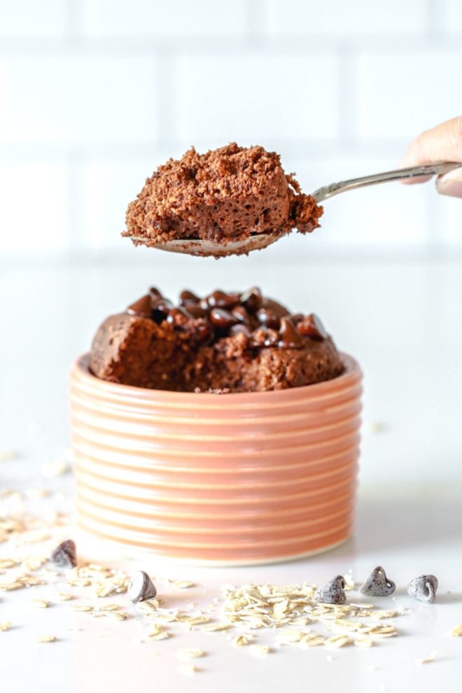 This is a side view of a hand holding a spoon with chocolate baked oatmeal above a small pink dish. Sticking out the top of the dish is a chocolate cake with chocolate chips on top.