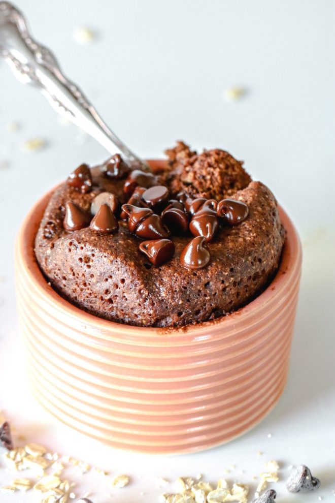 This is a side view looking at a light pink dish with chocolate baked oats inside. The baked oats are topped with melted chocolate chips and a spoon scooping out a bite. The dish sits on a white counter with oats scattered around.