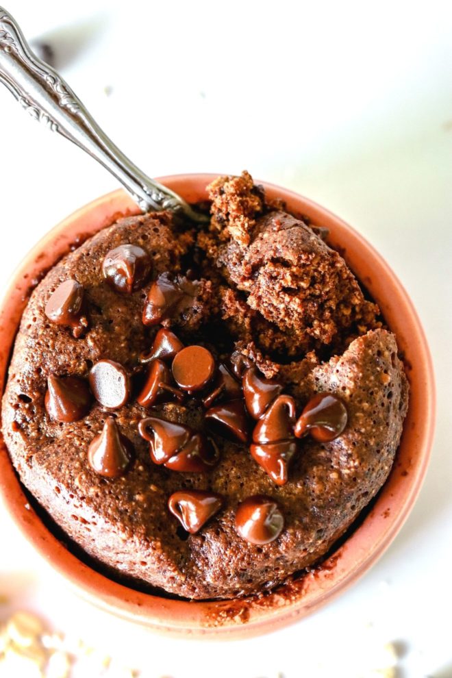 This is an overhead view looking into a light pink dish with chocolate baked oats inside. The baked oats are topped with melted chocolate chips and a spoon scooping out a bite. The dish sits on a white counter with oats scattered around.