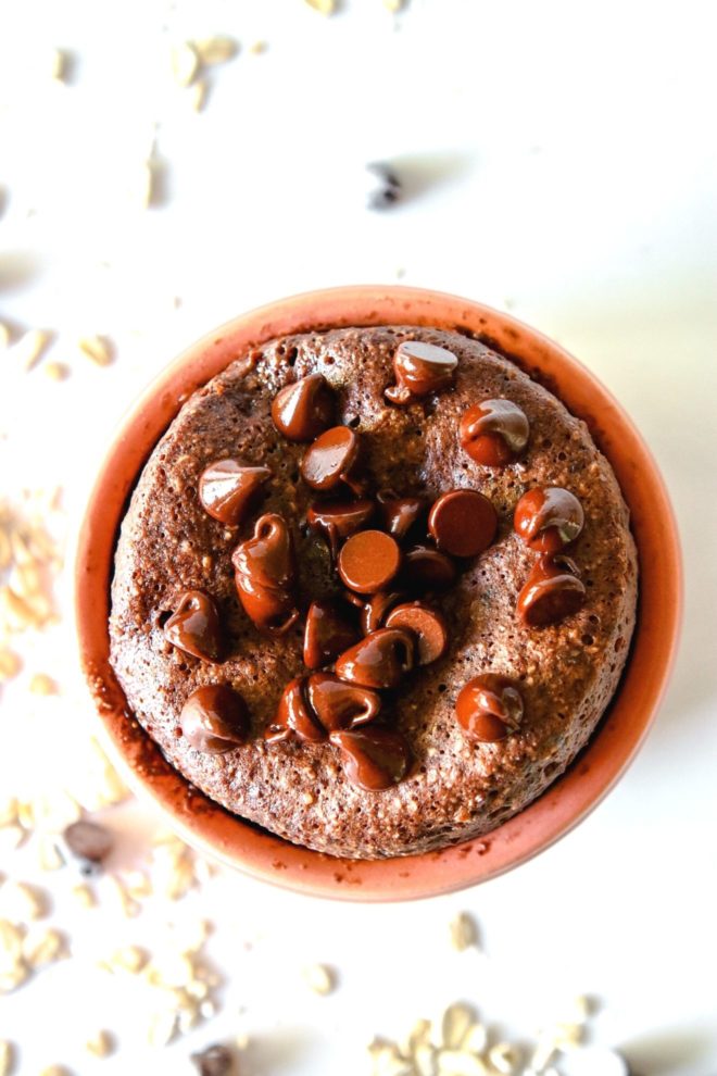 This is an overhead view looking into a light pink dish with chocolate baked oats inside. The baked oats are topped with melted chocolate chips. The dish sits on a white counter with oats and chocolate chips scattered around.