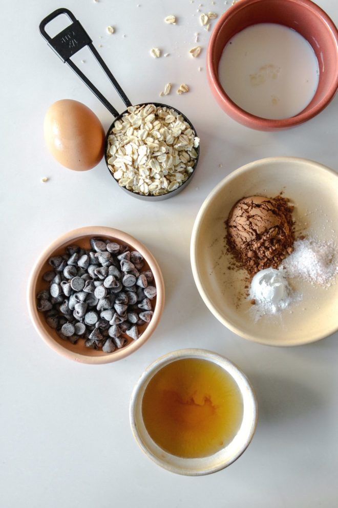 This is an overhead image looking at the ingredients to make chocolate baked oats. The ingredients are milk, oats, an egg, chocolate chips, cocoa powder, salt, baking powder, and agave nectar on a white counter.