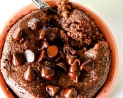 This is an overhead view looking into a light pink dish with chocolate baked oats inside. The baked oats are topped with melted chocolate chips and a spoon scooping out a bite. The dish sits on a white counter with oats scattered around. Text overlay reads "chocolate baked oats 1-min microwave."