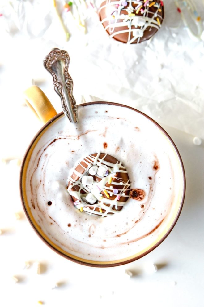 This is an overhead image of a yellow mug with foamy milk and a chocolate ball in the center. The chocolate sphere has sprinkles and marshmallows on top. The mug sits on a white surface with another chocolate ball to the top right of the image.