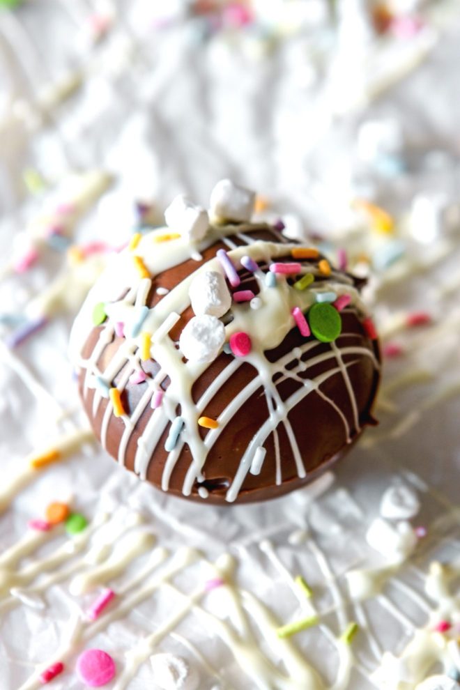 This is a closeup of a hot chocolate bomb drizzled with white chocolate, colorful sprinkles, and small marshmallows. The chocolate sphere sits on a white surface with more white chocolate drizzles and sprinkles around it.