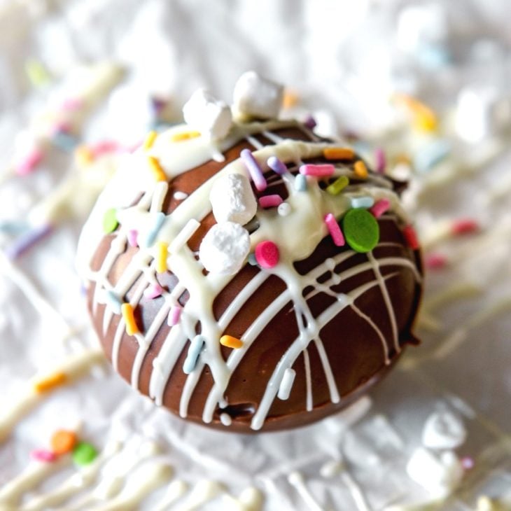 This is a closeup of a hot chocolate bomb drizzled with white chocolate, colorful sprinkles, and small marshmallows. The cocoa bomb sits on a white surface with more white chocolate drizzles and sprinkles around it.