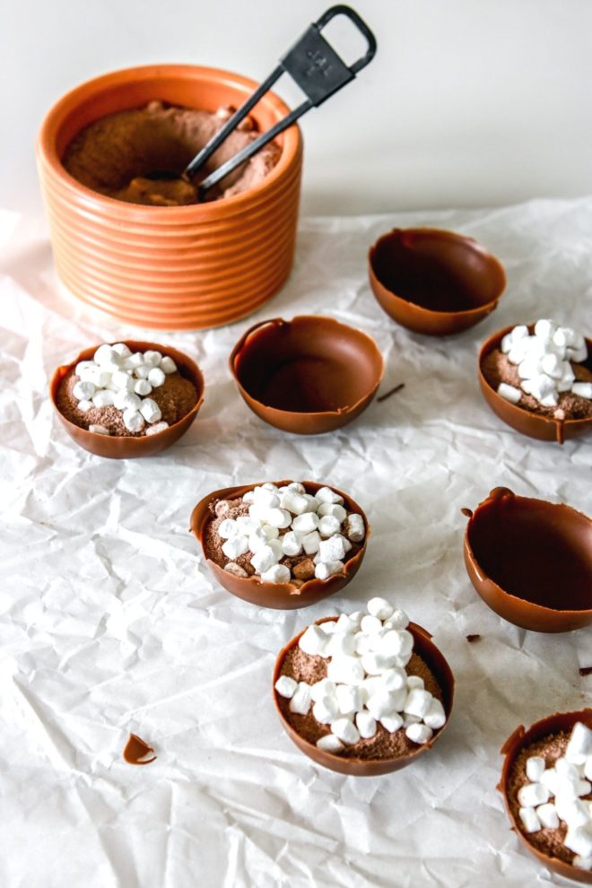 This is a side overhead angle image of half-sphere chocolate shells. Some are filled with hot chocolate mix and marshmallows and some are left empty. The chocolate shells sit on a white piece of parchment paper with a small bowl of hot cocoa mix is in the back of the image.