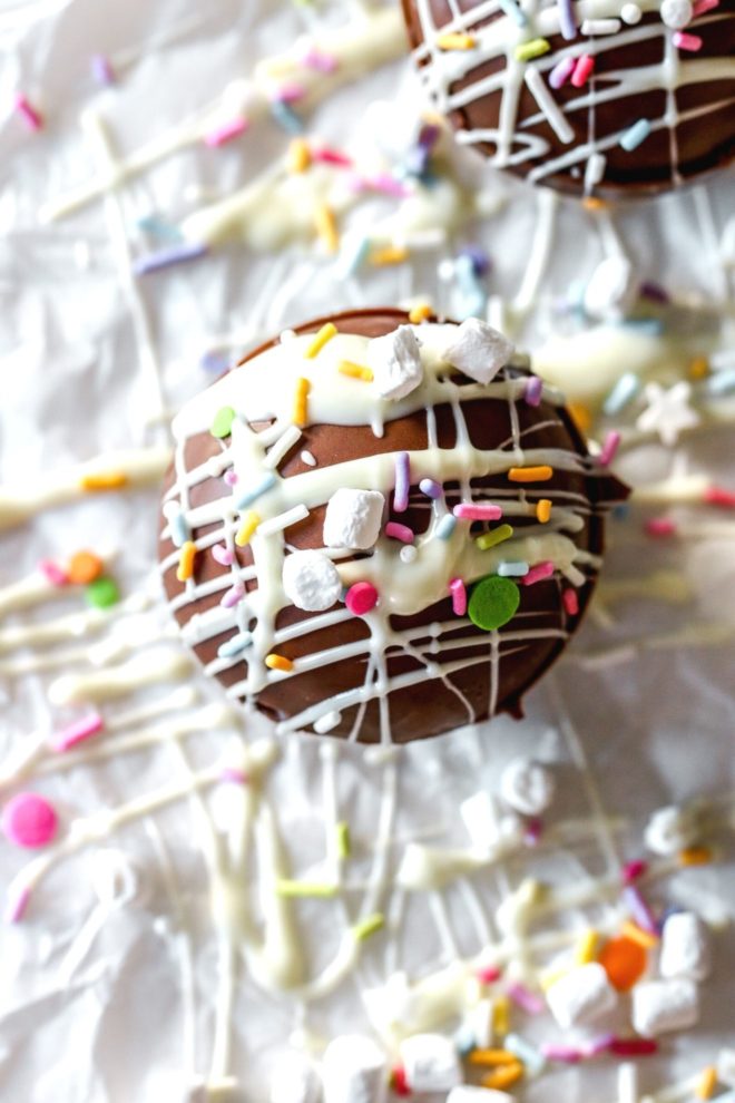 This is an overhead image of a chocolate sphere drizzled with white chocolate and topped with sprinkles and marshmallows. It sits on a white piece of parchment paper with another bomb to the top right corner of the image. There are more sprinkles on the white surface around the cocoa bombs.