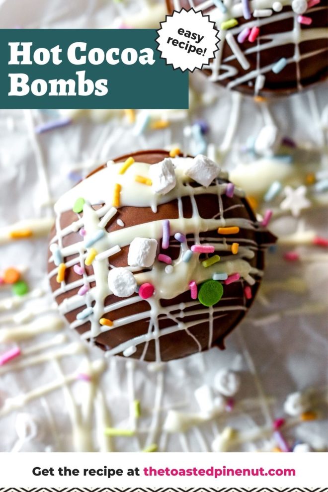 This is an overhead image of a chocolate sphere drizzled with white chocolate and topped with sprinkles and marshmallows. The cocoa bomb sits on a white piece of parchment paper with another bomb to the top right corner of the image. There are more sprinkles on the white surface around the cocoa bombs. Text overlay reads "hot cocoa bombs easy recipe!"