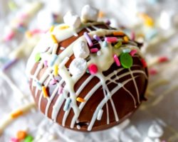 This is a closeup of a hot chocolate bomb drizzled with white chocolate, colorful sprinkles, and small marshmallows. The cocoa bomb sits on a white surface with more white chocolate drizzles and sprinkles around it. Text overlay reads "super fun hot cocoa bombs."