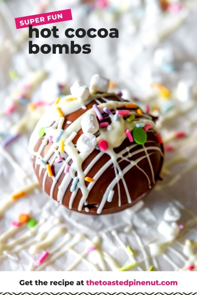 This is a closeup of a hot chocolate bomb drizzled with white chocolate, colorful sprinkles, and small marshmallows. The cocoa bomb sits on a white surface with more white chocolate drizzles and sprinkles around it. Text overlay reads "super fun hot cocoa bombs."