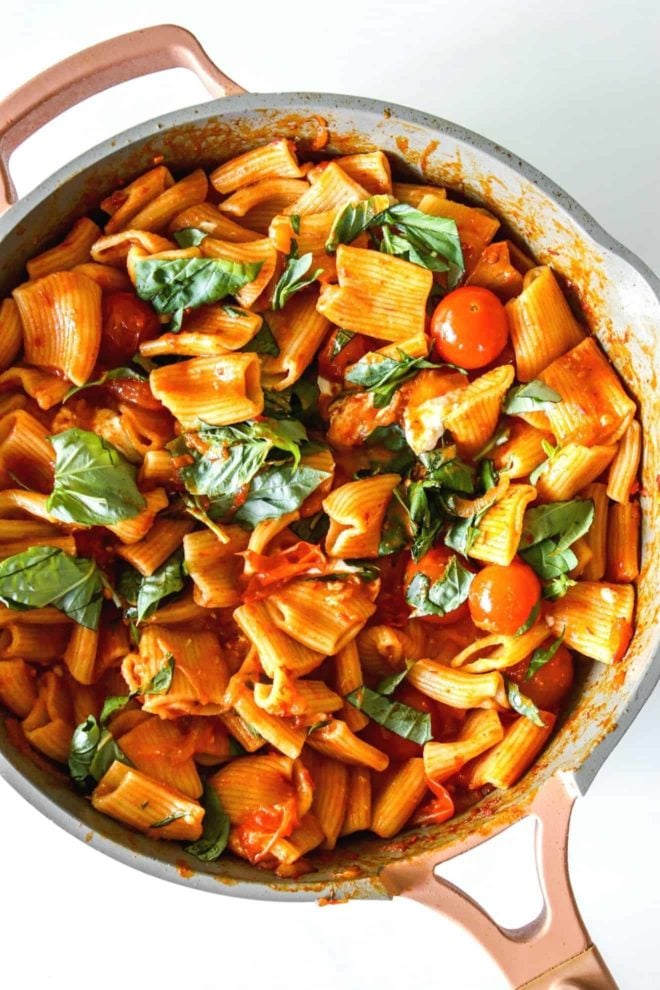 This is an overhead image of a pan with pasta in a harissa sauce, basil, and cherry tomatoes.