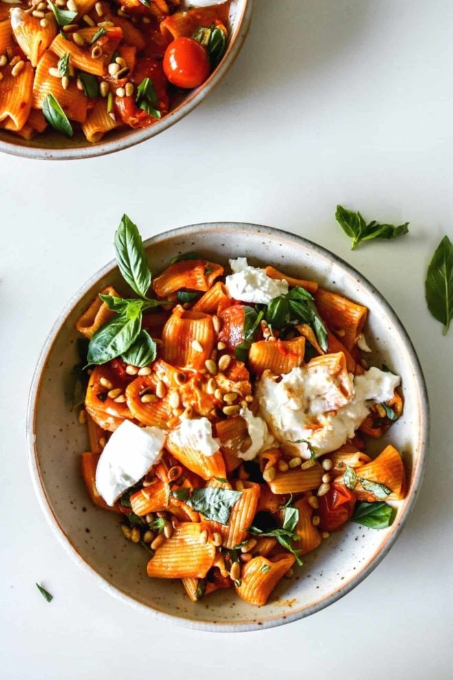 This is an overhead view of a bowl of harissa pasta. The pasta is topped with burrata, pine nuts, and basil.