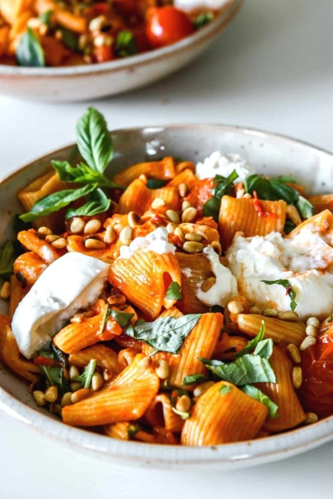 This is a side view of a bowl of harissa pasta. The pasta is topped with burrata, pine nuts, and basil.