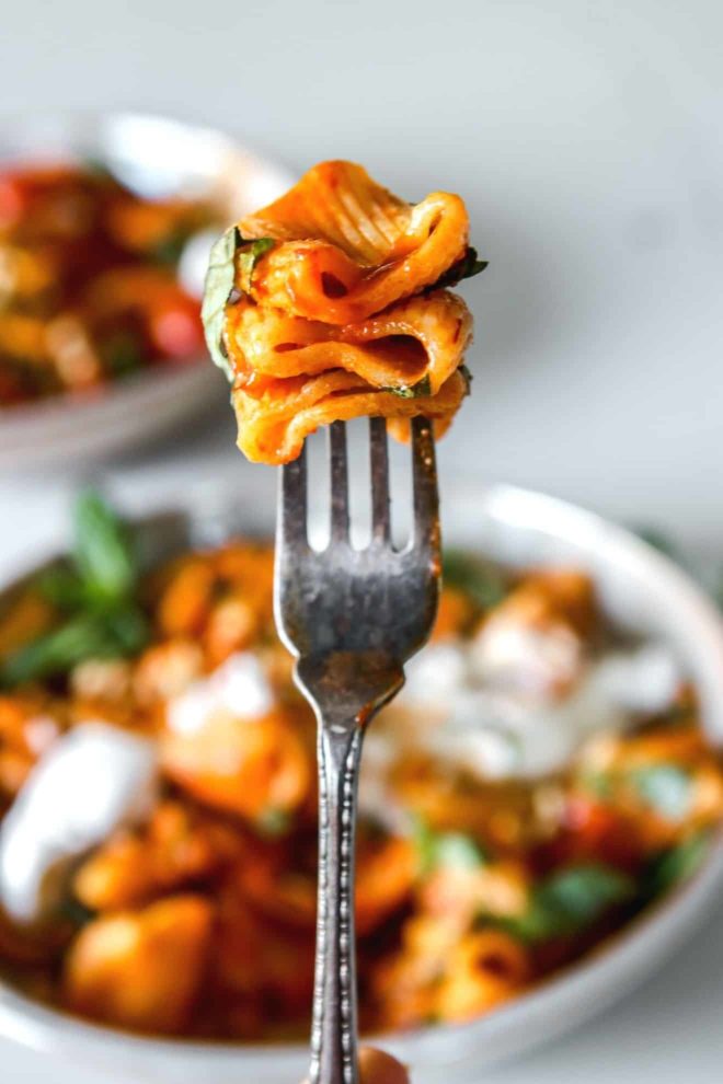 This is a closeup of a fork with three noodles on it. The pasta is in a red harissa sauce with some basil. Bowls of pasta are blurred in the background.