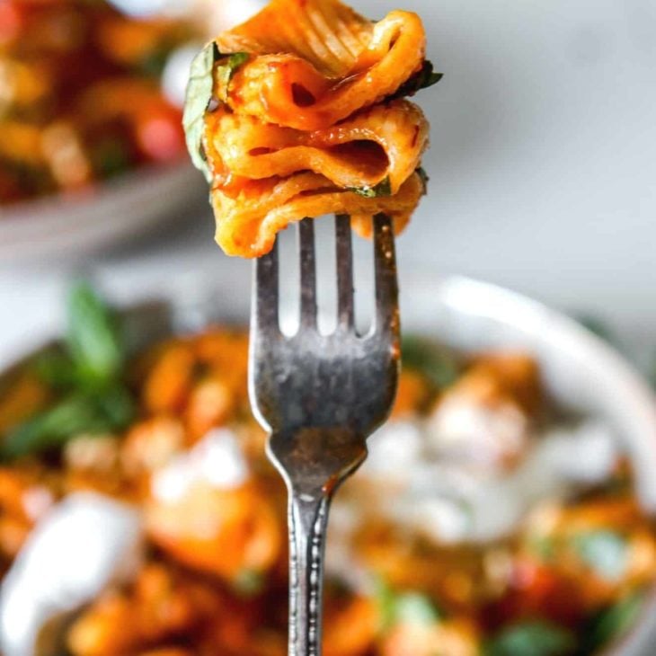 This is a closeup of a fork with three noodles on it. The pasta is in a red harissa sauce with some basil. Bowls of pasta are blurred in the background.