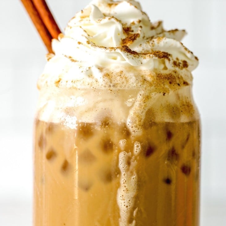 This is a closeup of a glass filled with ice, a latte, topped with whipped cream, pumpkin spice, and a cinnamon stick.