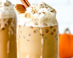 This is a side view of a glass with an iced pumpkin spice latte topped with whipped cream, a sprinkle of pumpkin spice, a cinnamon stick, and a waffle cookie. Another pumpkin spice latte is blurred in the background along with a small orange pumpkin. The glass sits on a white counter with pumpkin spice sprinkled on the counter along with some foam that has dripped down from the top of the glass. Text overlay reads "iced pumpkin spice latte get the recipe at thetoastedpinenut.com"