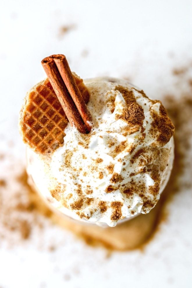 This is an overhead image of a latte topped with whipped cream, pumpkin spice, a cinnamon stick, and a waffle cookie.