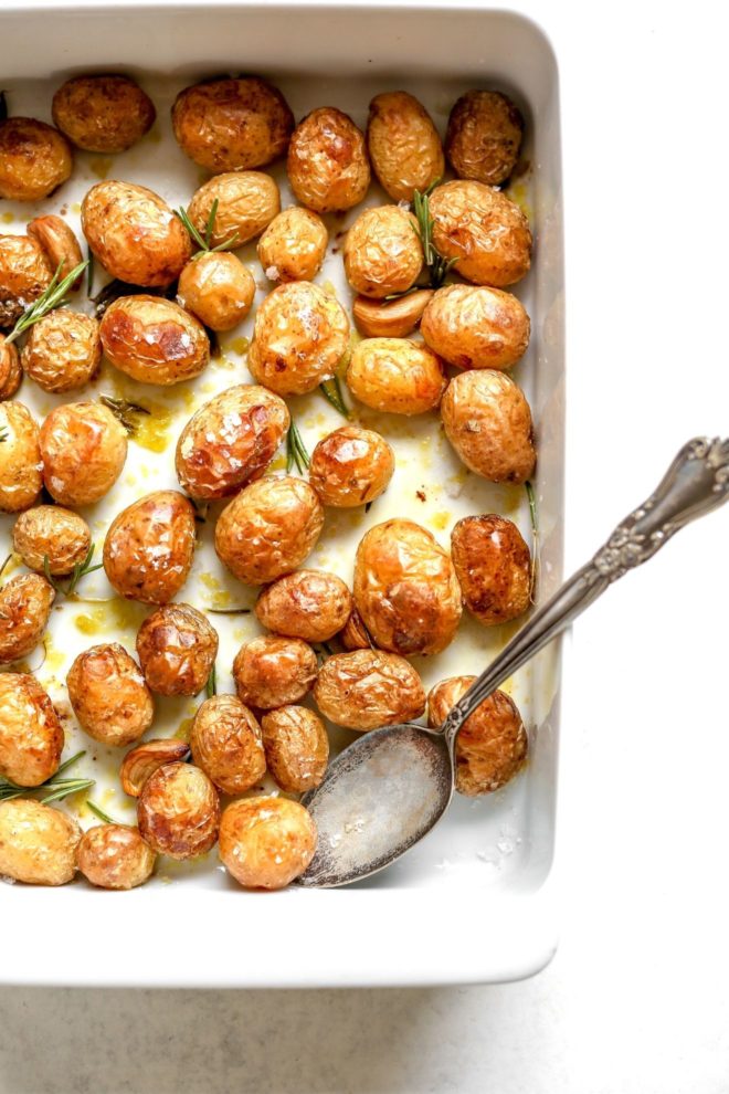 This is an overhead image of a white baking dish on a light grey surface. In the baking dish are roasted mini potatoes drizzled with oil and rosemary sprigs. A silver antique spoon is in the dish and leaning on the right side.