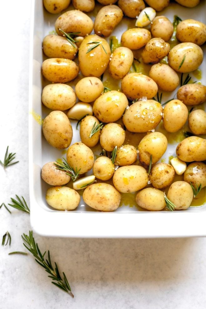 This is an overhead image of a white baking dish on a light grey surface. In the baking dish are raw mini potatoes drizzled with oil and rosemary sprigs. More rosemary sprigs are on the surface to the bottom left of the image.