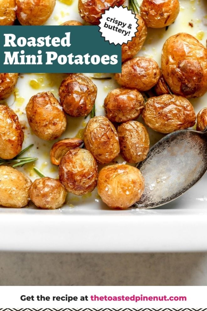This is an overhead image of a white baking dish on a light grey surface. In the baking dish are roasted mini potatoes drizzled with oil and rosemary sprigs. A silver antique spoon is in the dish and leaning on the right side. Text overlay reads "crispy & buttery roasted mini potatoes."