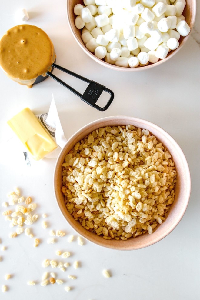 This is an overhead image of a bowl of rice krispies, a bowl of marshmallows, peanut butter, and butter on a white counter.