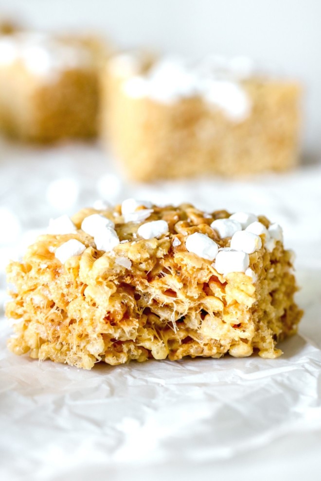 This is a side view of a peanut butter rice krispie treat with a bite taken out of it. The treat sits on a crumpled piece of white parchment paper with more treats blurred in the background.
