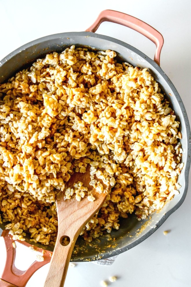 This is an overhead image of a pan with rice krispie treat mixture and a wooden spatula. The pan sits on a white background.