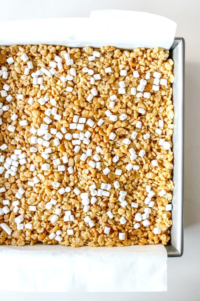 This is an overhead image of a pan with parchment paper with rice krispie treats pressed into the pan. The treats are topped with mini marshmallows.