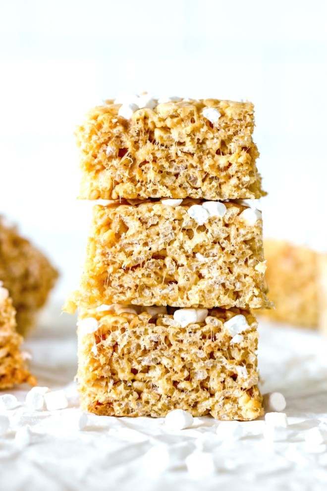 This is a side view of a stack of three rice krispie treats. The stack is on a white surface with more treats blurred in the background.