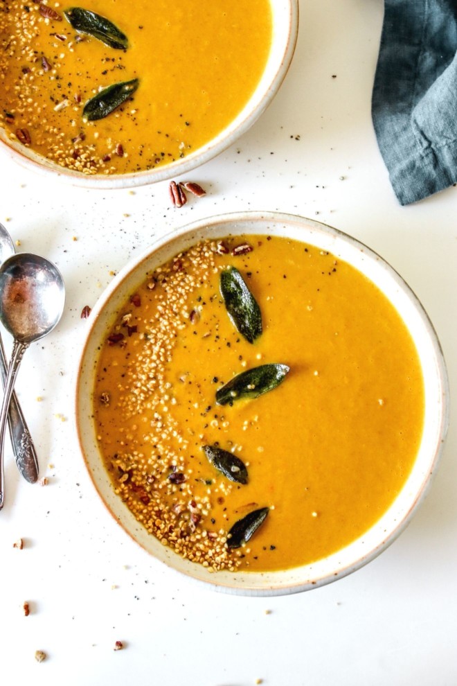 This is an overhead image of two shallow bowls of pumpkin soup. The soup is topped with fried sage leaves, pecans, and quinoa. The bowls sit on a white counter with antique spoons to the left of the bowl and a navy tea towel in the top right corner of the image.