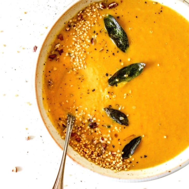 This is an overhead image of a shallow bowl with pumpkin soup on a white counter. The soup is topped with sage leaves, pecans, and quinoa. A spoon is dipping into the soup to the left side of the bowl. Another bowl of soup is to the top left of the image.