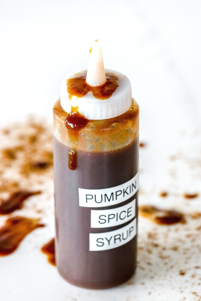 This is a side view of a plastic squeeze bottle with a deep caramel colored syrup inside. A white label reads "pumpkin spice syrup." The bottle sits on a white counter with pumpkin spice and some syrup drips around it.
