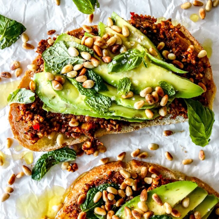This is an overhead image of two slices of avocado toast. The toast is topped with a harissa pesto and slices of avocado, toasted pine nuts, and herbs. The toast lays on a crinkled white piece of parchment paper.