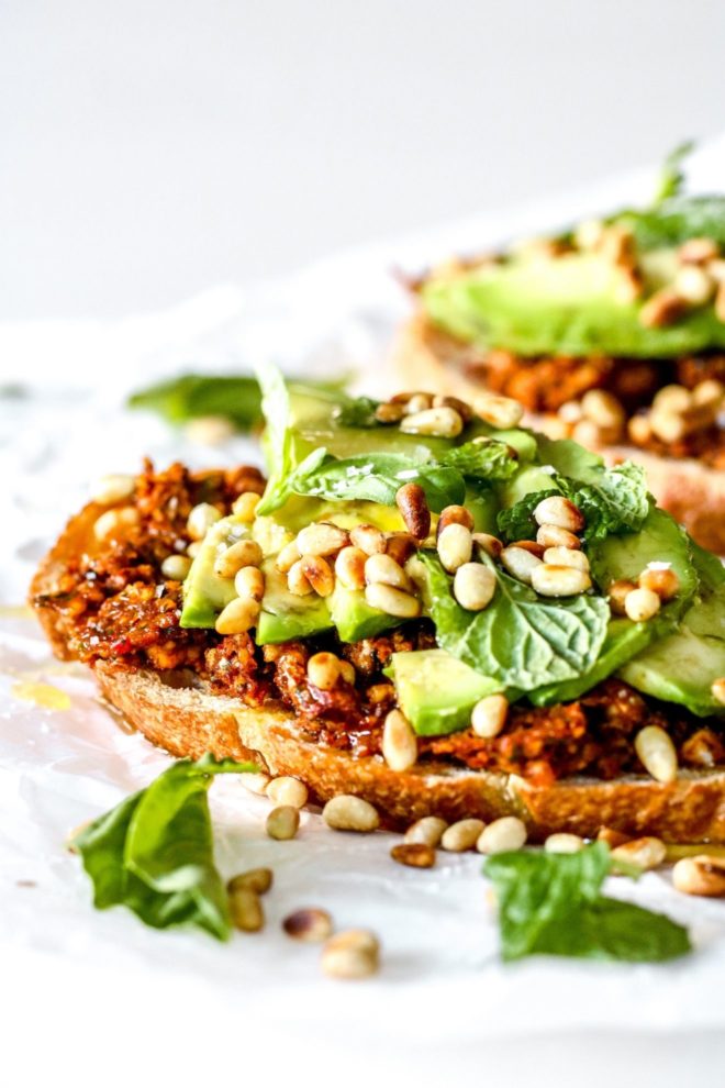 This is a side view of a slice of toast topped with harissa pesto, avocado, fresh herbs, and toasted pine nuts. The piece of toast sits on a white piece of parchment paper with another avocado toast blurred in the background.