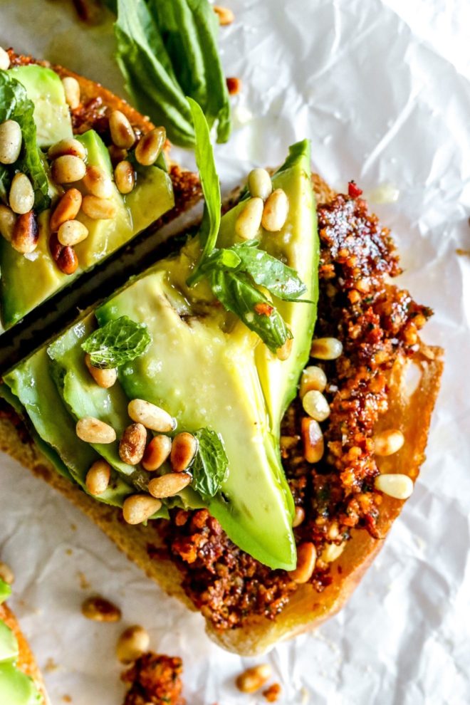 This is an overhead image of a slice of avocado toast cut in half. The toast is topped with a harissa pesto and slices of avocado, toasted pine nuts, and herbs. The toast lays on a crinkled white piece of parchment paper.