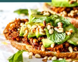 This is a side view of a slice of toast topped with harissa pesto, avocado, fresh herbs, and toasted pine nuts. The piece of toast sits on a white piece of parchment paper with another avocado toast blurred in the background. Text overlay reads "the best avocado toast vegan!"