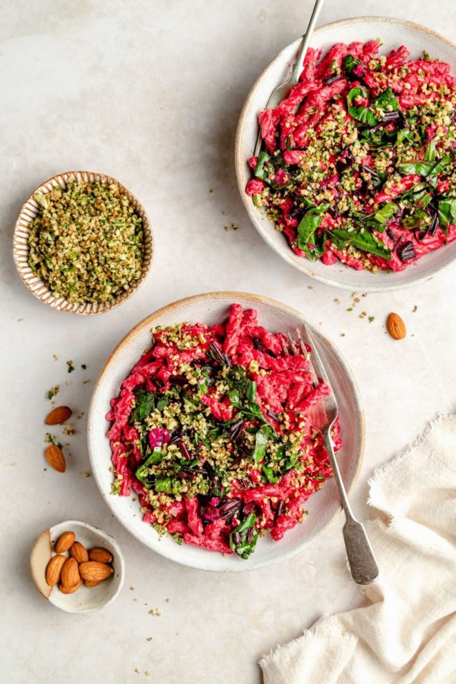 This is an overhead image of two dishes on a light surface. In the dishes are hot pink beetroot pastas topped with sautéed greens and chopped nuts. A small bowl of almonds and a small bowl of gremolata are to the left of the bowl.