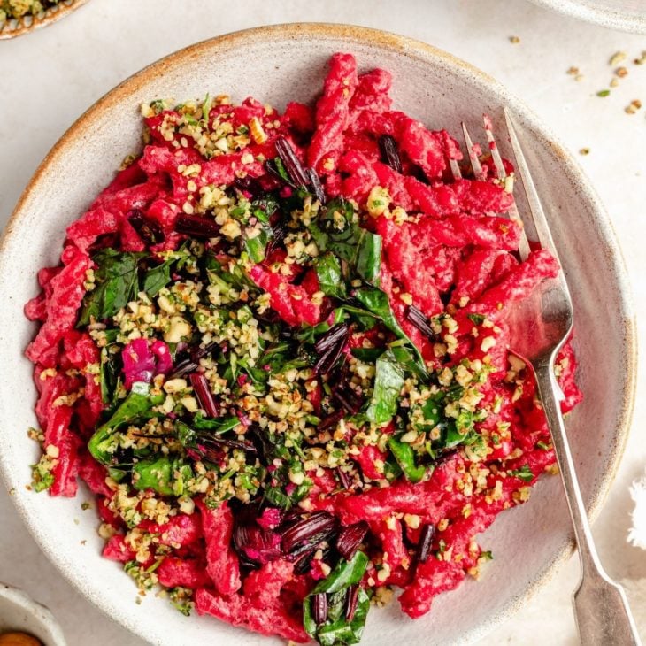 This is an overhead image of a shallow bowl with hot pink beetroot pasta topped with sautéed greens and a nutty mixture on top. A fork dips into the bowl and leans against the right side of the dish. Another bowl with pasta is to the top right corner of the image. A small bowl with nutty gremolata is to the top left corner of the image.