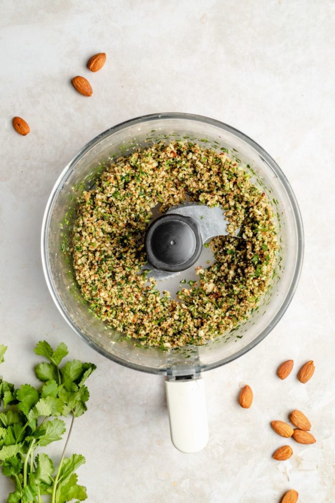 This is an overhead image of a food processor with nuts and herbs chopped up inside it. The food processor sits on a white surface with whole almonds to the top left corner and bottom right corner. Cilantro leaves are to the bottom left of the image.