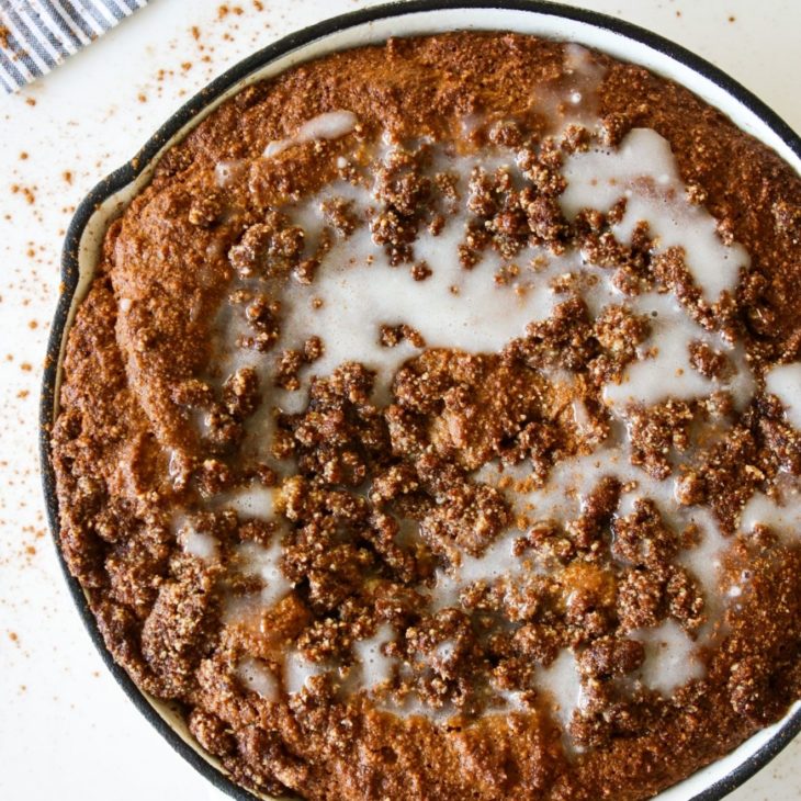 This is an overhead image of a white skillet with a coffee cake topped with a crumble streusel and vanilla glaze icing. The skillet sits on a white counter and a grey and white pinstripe tea towel is to the top left corner. The skillet and counter is sprinkled with cinnamon.